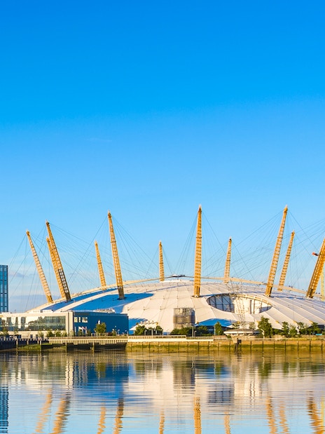 O2 Arena in London with surrounding buildings and cranes reflected in the Thames River.