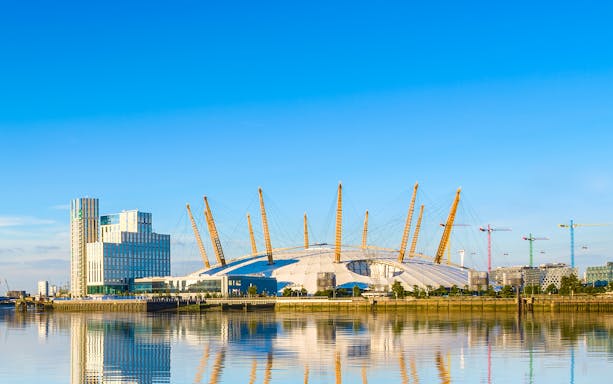 O2 Arena in London with surrounding buildings and cranes reflected in the Thames River.