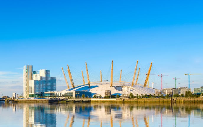 O2 Arena in London with surrounding buildings and cranes reflected in the Thames River.
