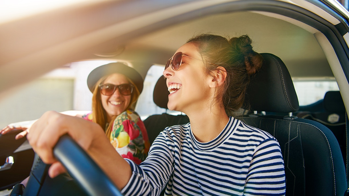 Woman driving car on road trip with friend smiling in passenger seat.