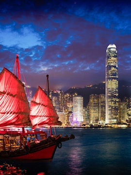 Red-sailed Aqua Luna cruise ship in Victoria Harbour, Hong Kong skyline at dusk.
