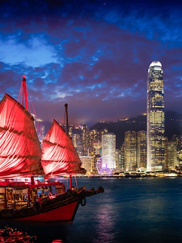 Red-sailed Aqua Luna cruise ship in Victoria Harbour, Hong Kong skyline at dusk.