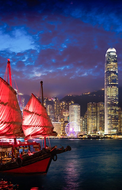 Red-sailed Aqua Luna cruise ship in Victoria Harbour, Hong Kong skyline at dusk.