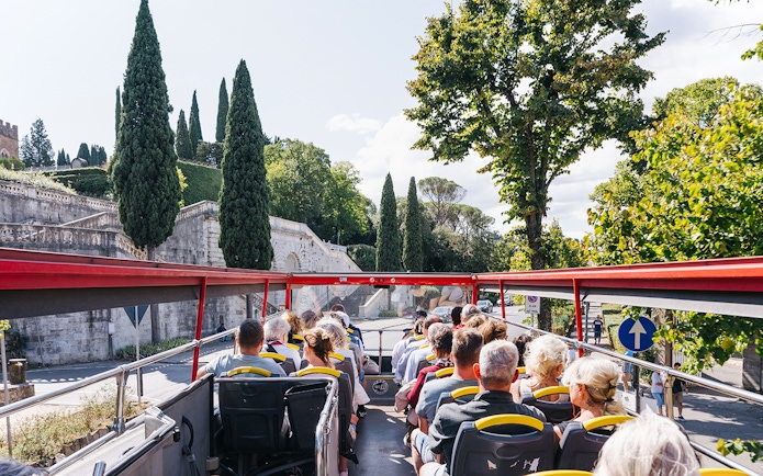Open-top bus with tourists on Florence Hop-On Hop-Off tour passing historic stone wall and cypress trees.