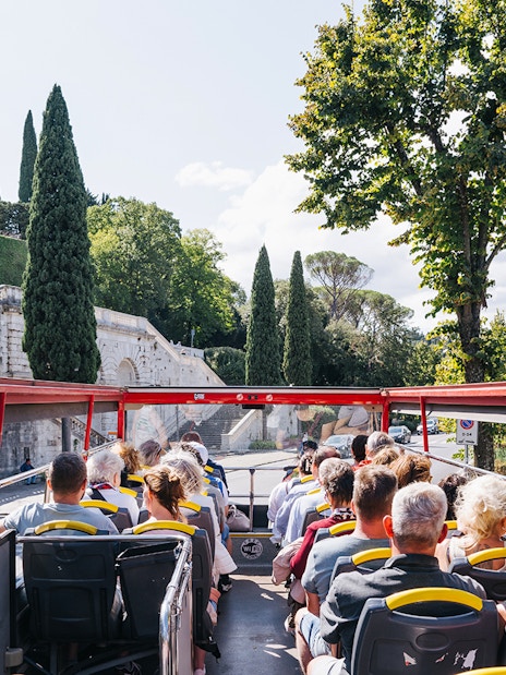 Open-top bus with tourists on Florence Hop-On Hop-Off tour passing historic stone wall and cypress trees.
