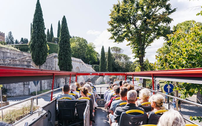 Open-top bus with tourists on Florence Hop-On Hop-Off tour passing historic stone wall and cypress trees.