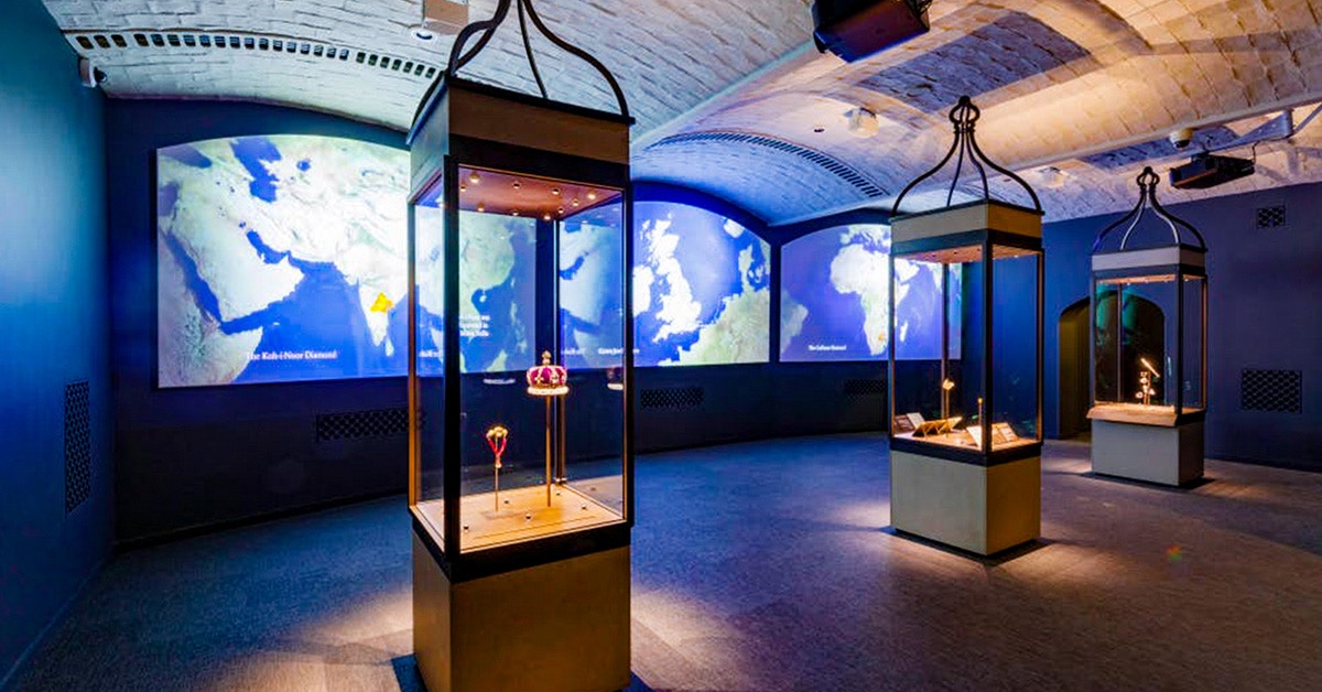 Crown jewels display at Tower of London with illuminated maps in the background.