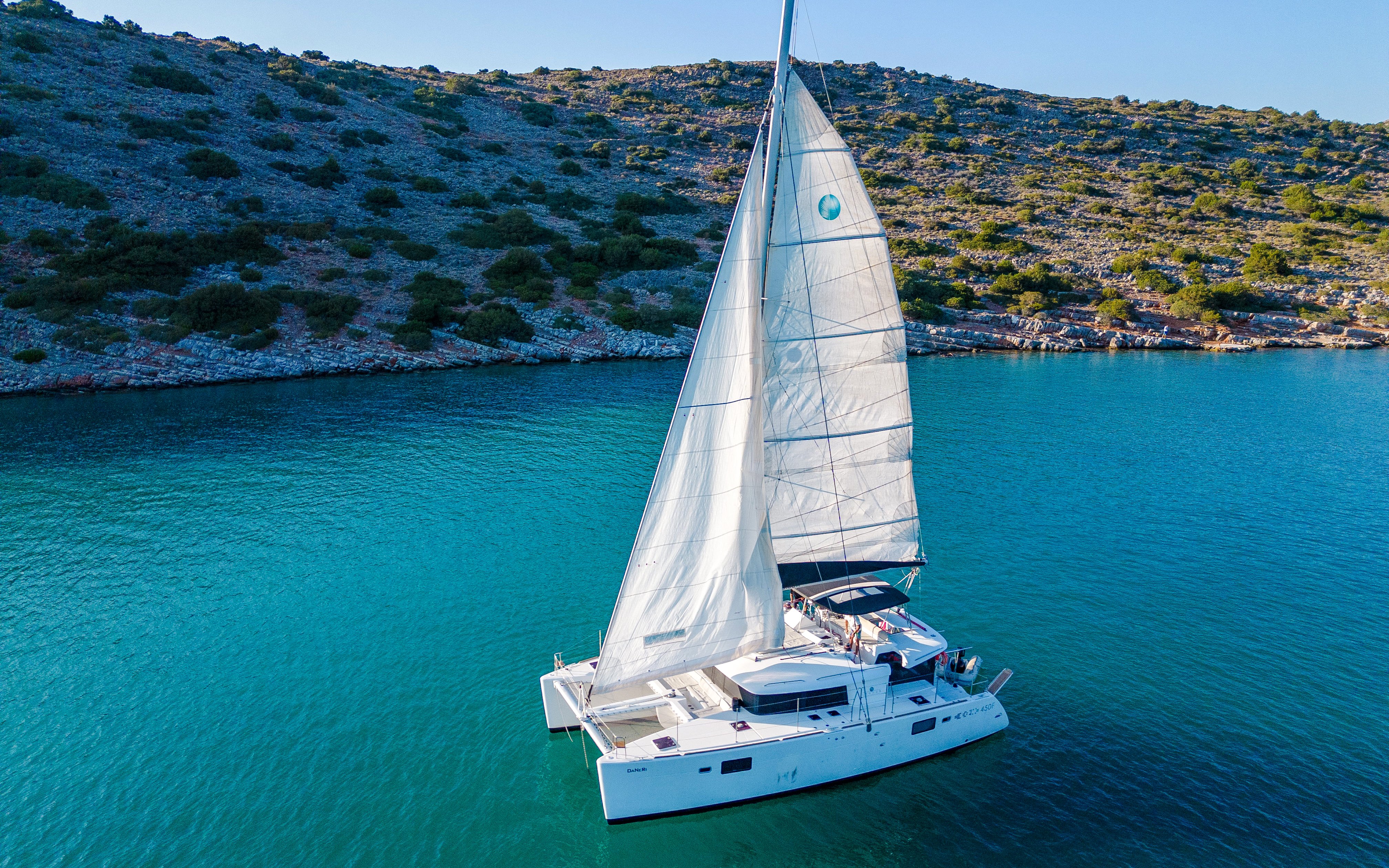 Catamaran sailing near Spinalonga, Crete, with rocky coastline in the background.