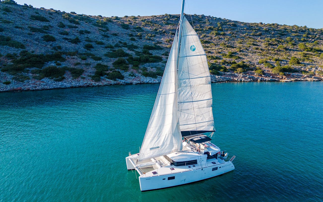 Catamaran sailing near Spinalonga, Crete, with rocky coastline in the background.