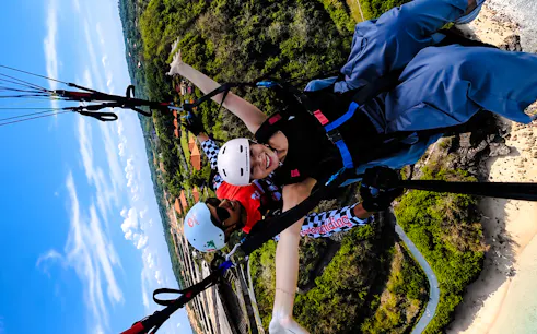 Tandem paragliding over Bali coastline with lush greenery and beach below.