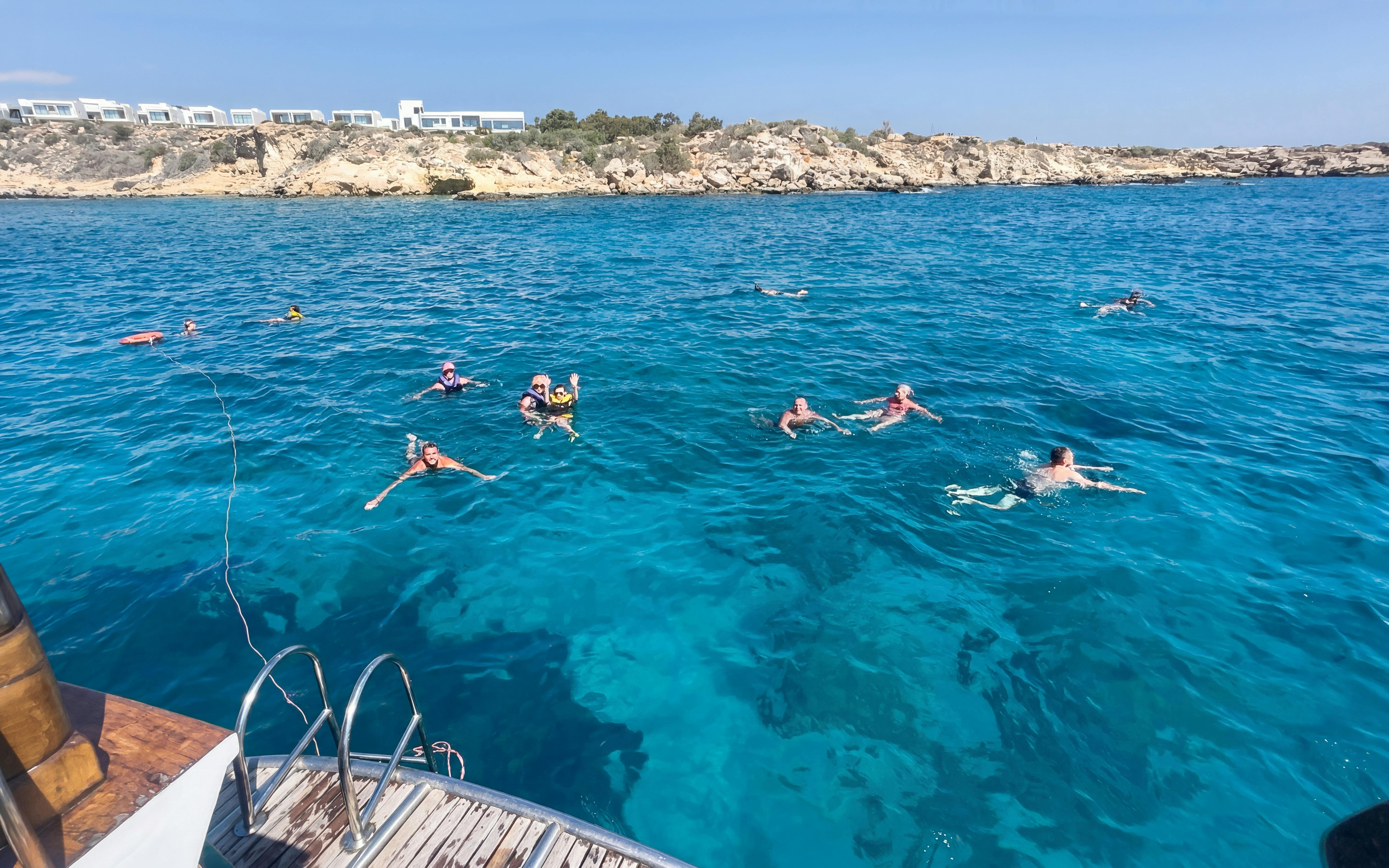 People swimming near a boat in the Blue Lagoon, Protaras, with rocky coastline in the background.