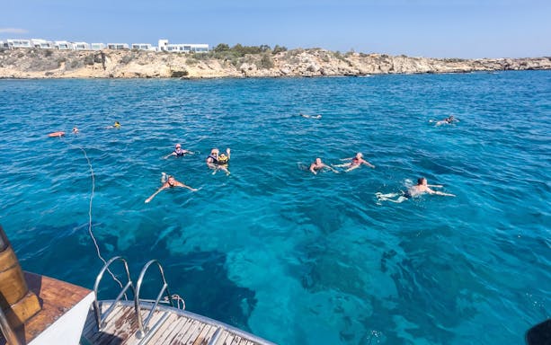People swimming near a boat in the Blue Lagoon, Protaras, with rocky coastline in the background.