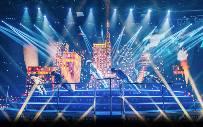 Performers on stage at Coco Bongo Disco with cityscape backdrop and vibrant lights.