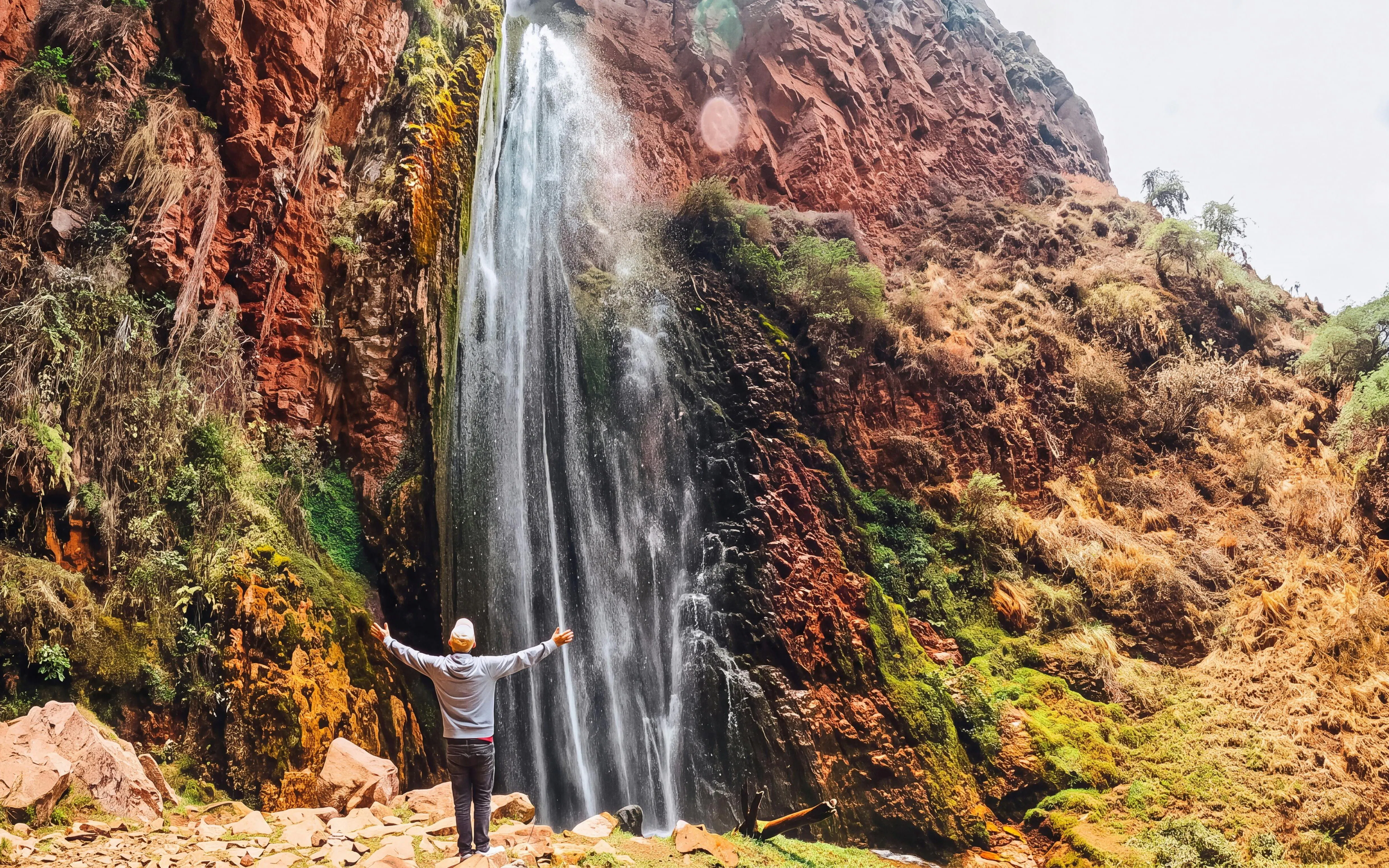 Person standing with arms outstretched in front of Perolniyoc Waterfall, Sacred Valley, Peru.