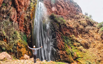 Person standing with arms outstretched in front of Perolniyoc Waterfall, Sacred Valley, Peru.