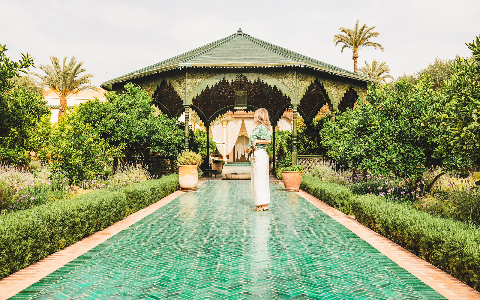 Woman standing on a green tiled path in Jardin Secret, surrounded by lush greenery and a pavilion.
