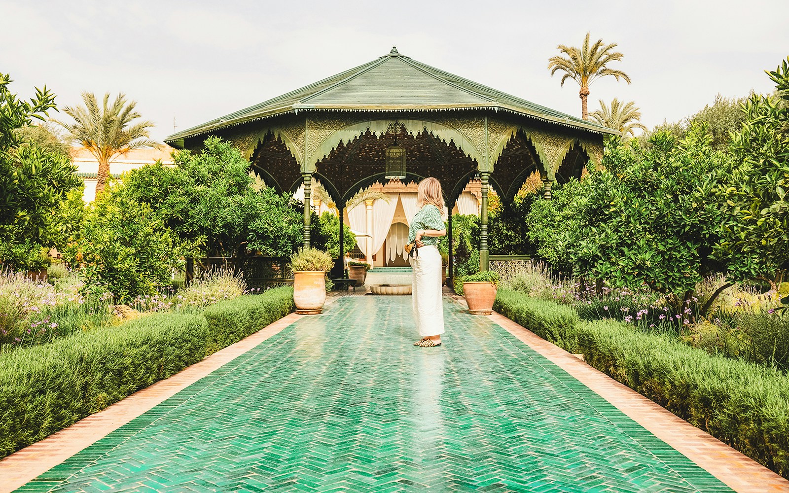 Woman standing on a green tiled path in Jardin Secret, surrounded by lush greenery and a pavilion.