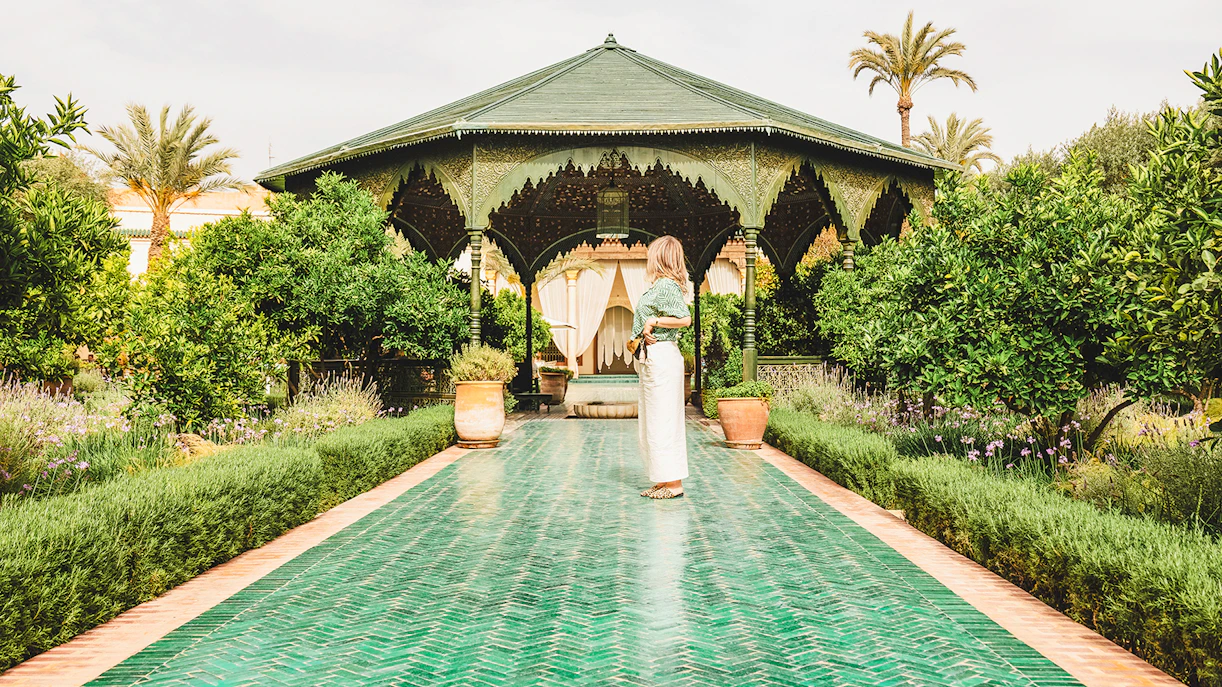 Woman standing on a green tiled path in Jardin Secret, surrounded by lush greenery and a pavilion.