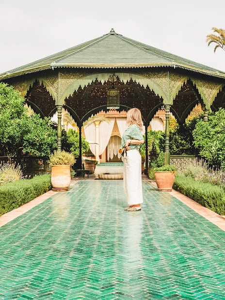Woman standing on a green tiled path in Jardin Secret, surrounded by lush greenery and a pavilion.