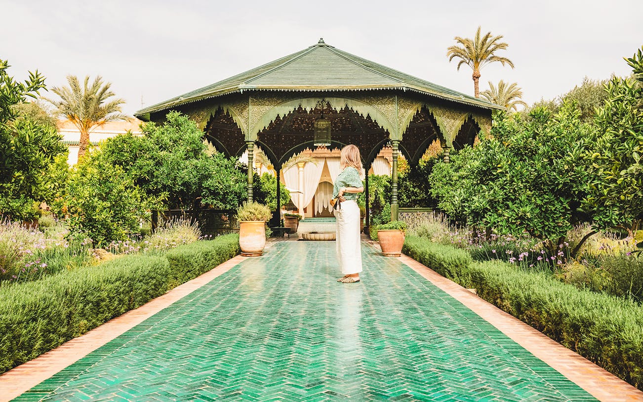 Woman standing on a green tiled path in Jardin Secret, surrounded by lush greenery and a pavilion.