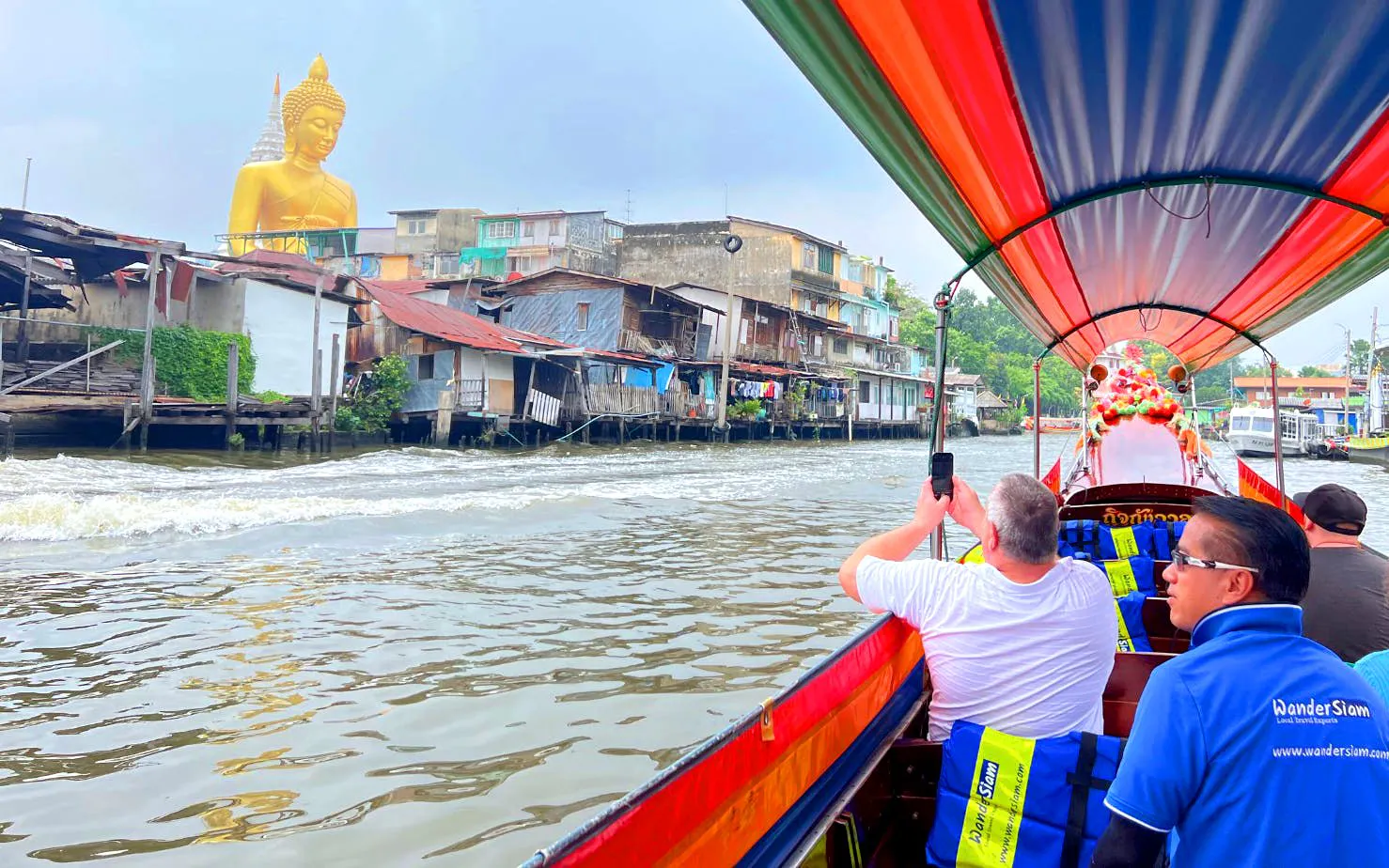 Longtail boat on Bangkok canal passing houses and a large Buddha statue.