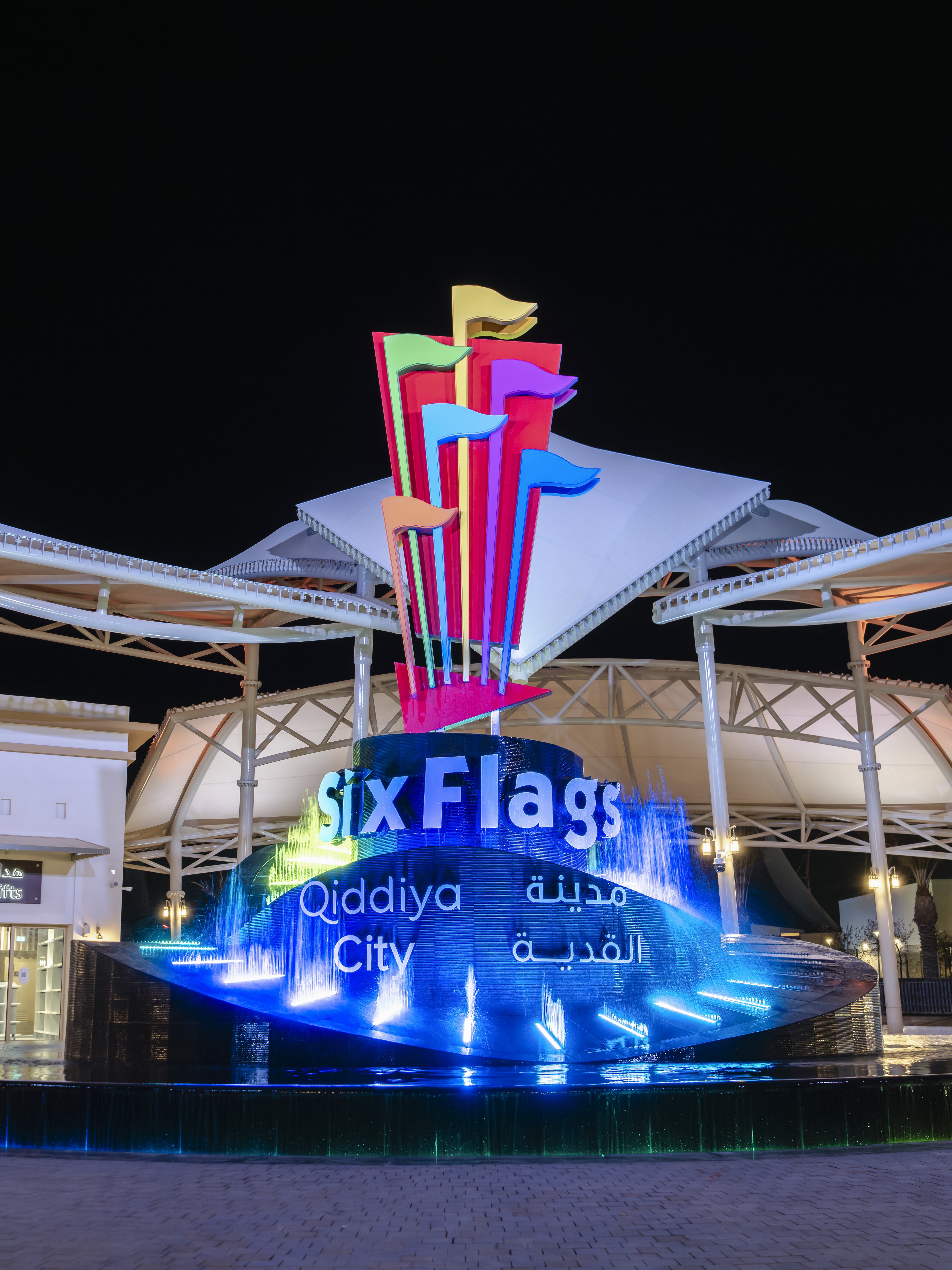 Six Flags Qiddiya City entrance with illuminated fountain and colorful flags at night.