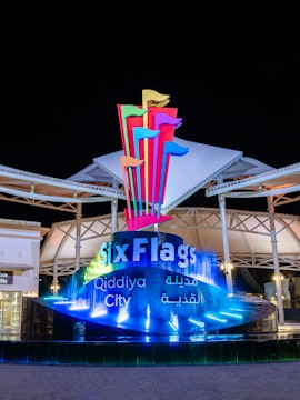 Six Flags Qiddiya City entrance with illuminated fountain and colorful flags at night.