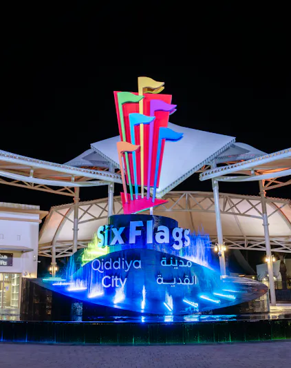 Six Flags Qiddiya City entrance with illuminated fountain and colorful flags at night.