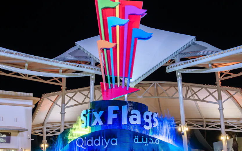 Six Flags Qiddiya City entrance with illuminated fountain and colorful flags at night.