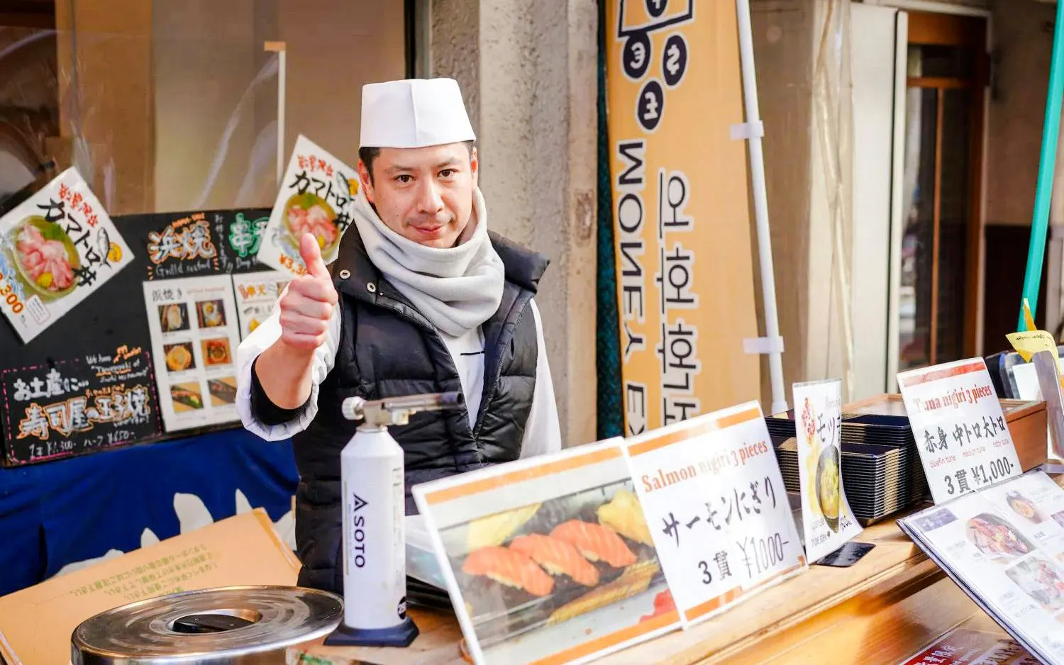 Shopkeeper giving thumbs up at Tsukiji Fish Market stall with sushi menu signs.