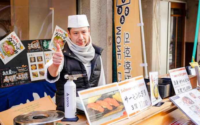 Shopkeeper giving thumbs up at Tsukiji Fish Market stall with sushi menu signs.