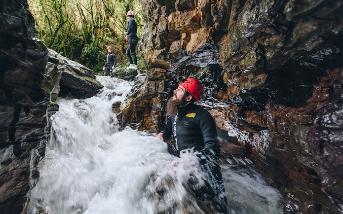 Adventurers in wetsuits and helmets navigate rocky stream in Waitomo cave.