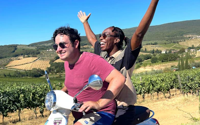Two people riding a Vespa through Chianti vineyards during a sunset winery tour.