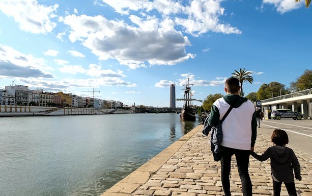 Father and child walking along the Guadalquivir River in Triana, Seville, with a historic ship in view.