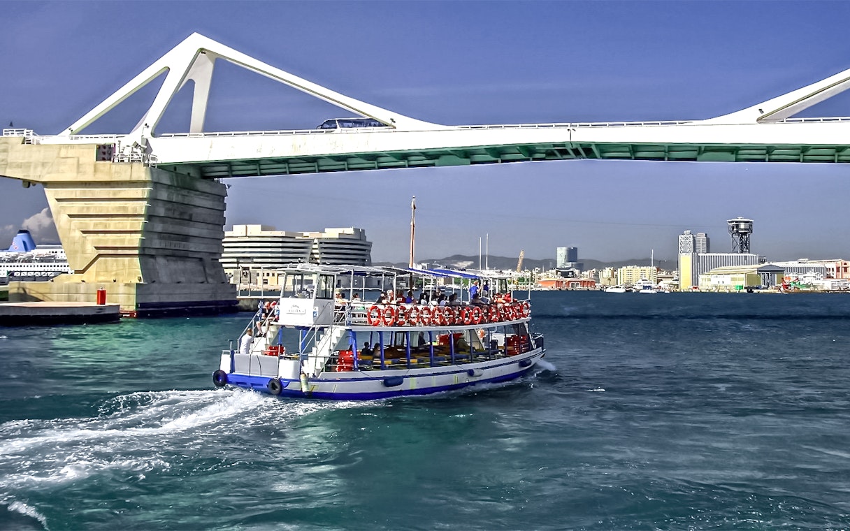 Las Golondrinas boat passing under a bridge in Barcelona harbor.