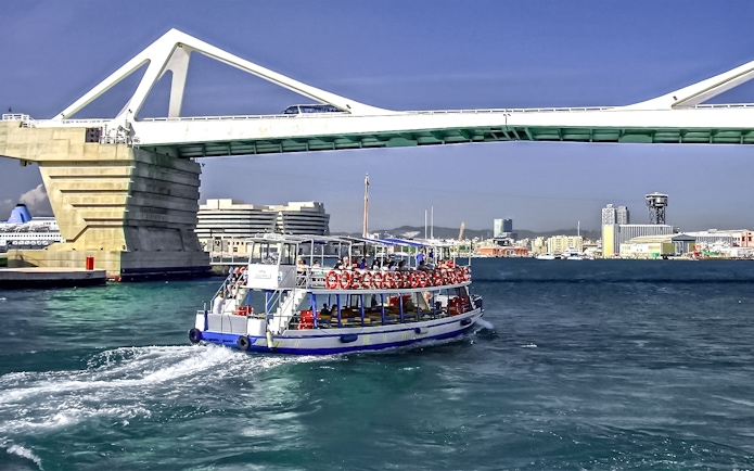 Las Golondrinas boat passing under a bridge in Barcelona harbor.
