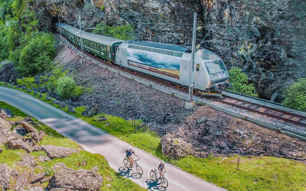 Flåm Railway train passing through rocky landscape with cyclists on nearby path.