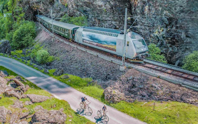 Flåm Railway train passing through rocky landscape with cyclists on nearby path.