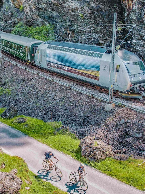 Flåm Railway train passing through rocky landscape with cyclists on nearby path.