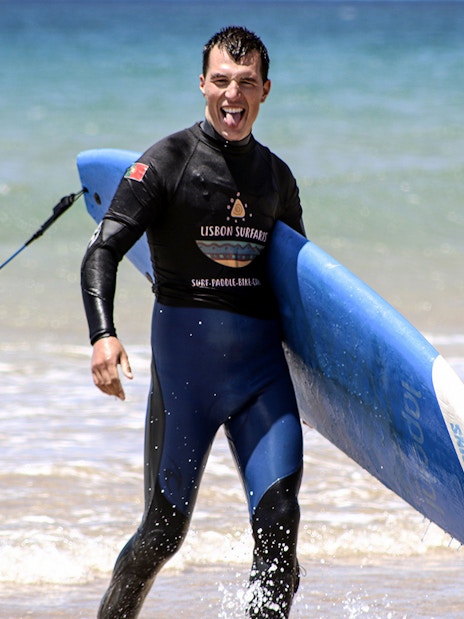 Surfer carrying a blue surfboard on a beach during a group surf lesson in Lisbon.