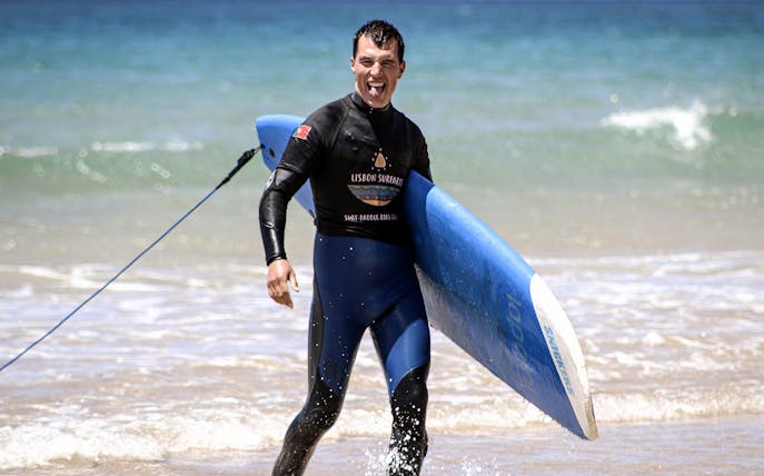 Surfer carrying a blue surfboard on a beach during a group surf lesson in Lisbon.