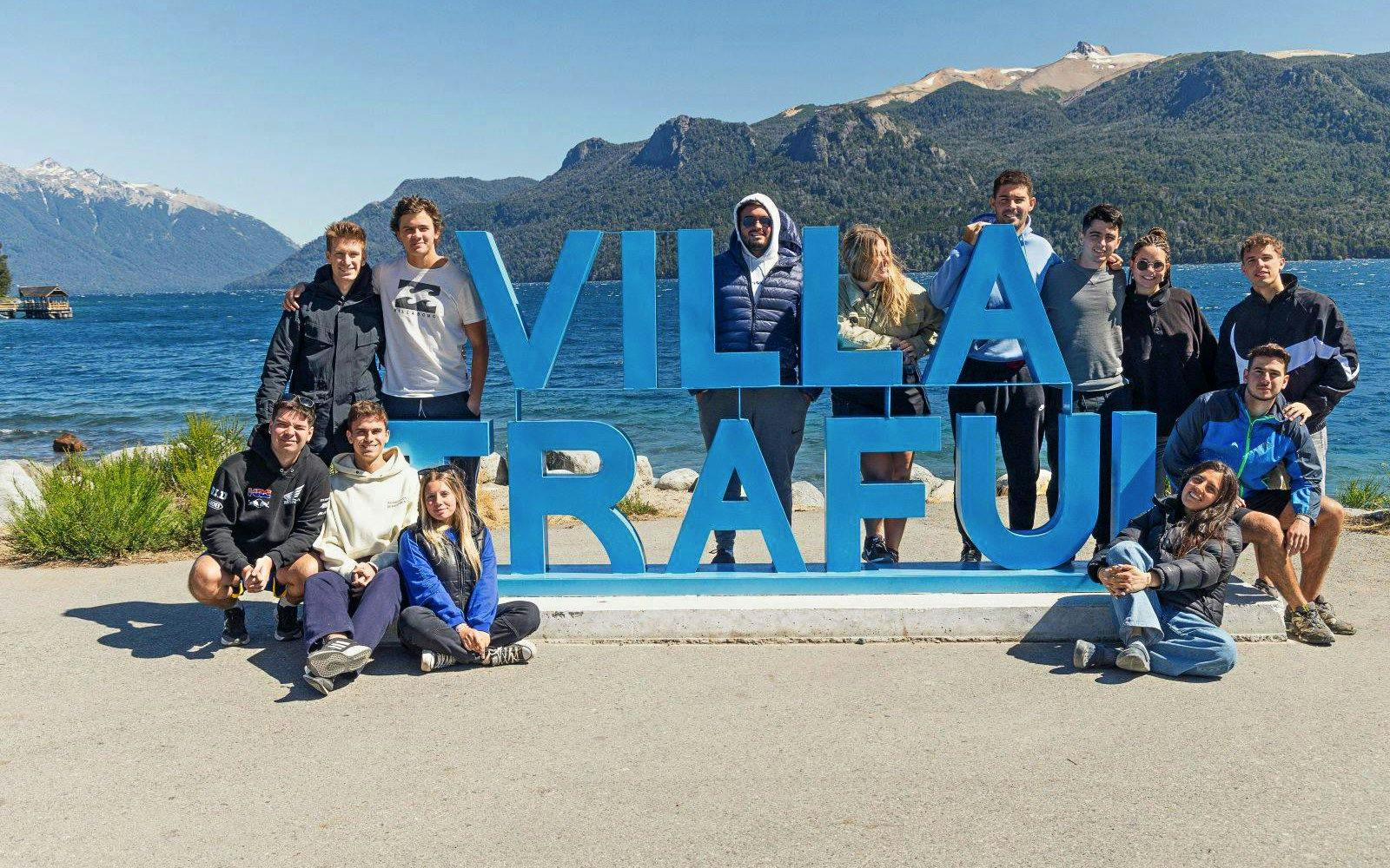Travelers gathered at Villa Traful sign with Patagonian mountains and lake in the background.