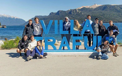 Travelers gathered at Villa Traful sign with Patagonian mountains and lake in the background.