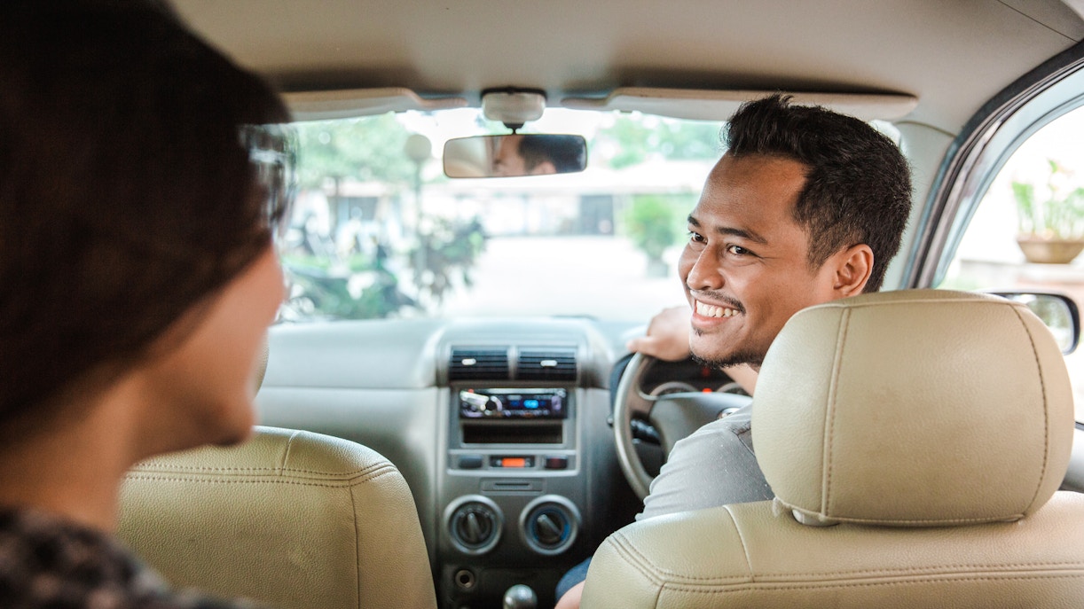 Person smiling while driving a taxi with a passenger in the back seat.
