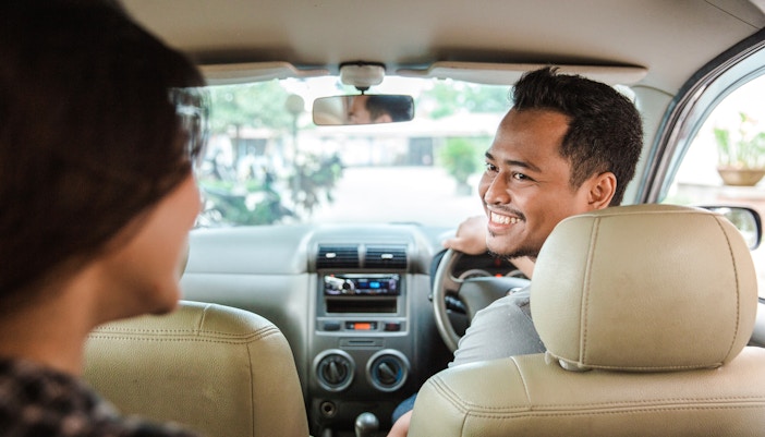 Person smiling while driving a taxi with a passenger in the back seat.