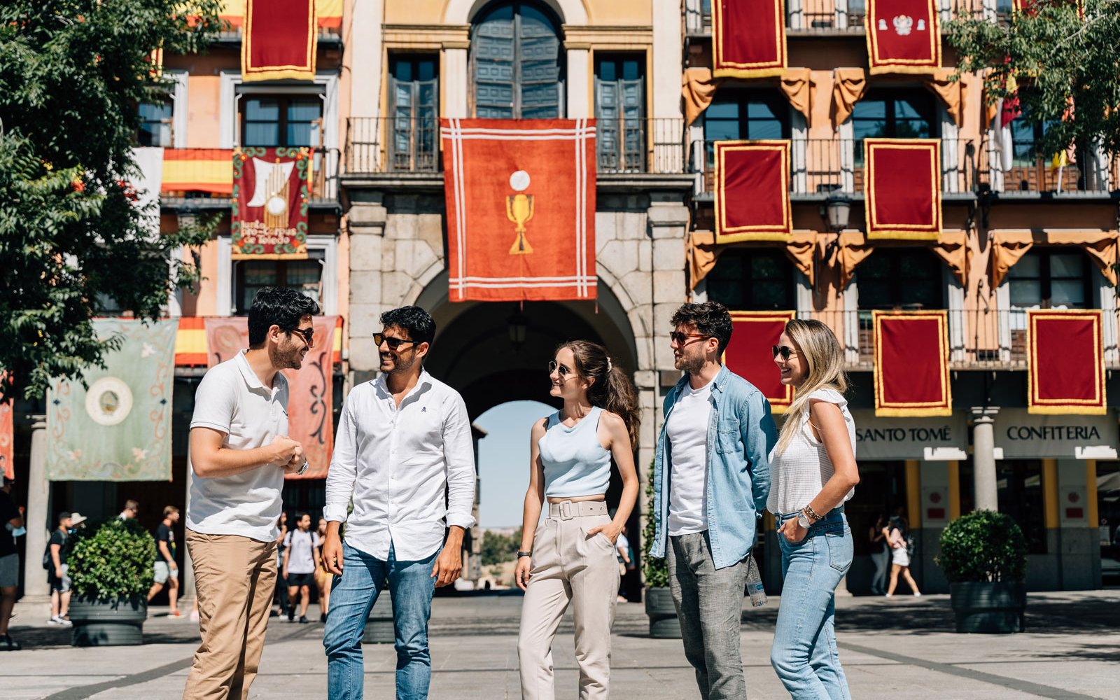 Tourists with guide at Corpus Christi procession in Toledo, Spain, standing in front of decorated building.