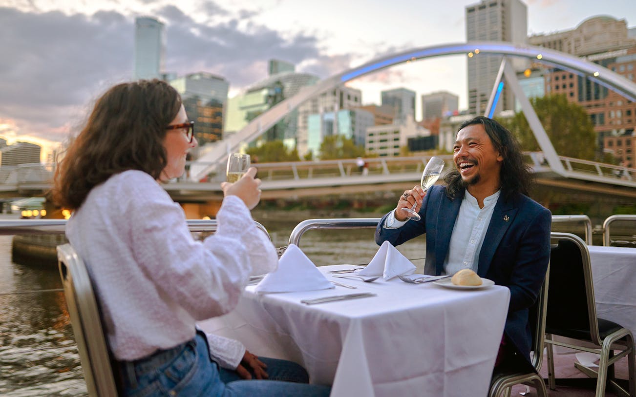 Couple enjoying wine on the Spirit of Melbourne Dinner Cruise with city skyline in the background.