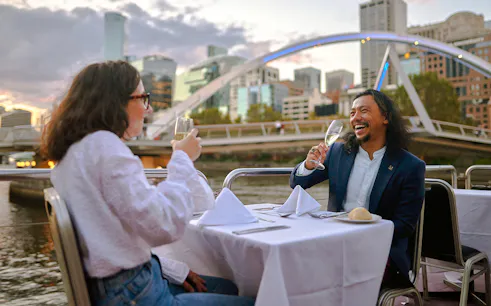 Couple enjoying wine on the Spirit of Melbourne Dinner Cruise with city skyline in the background.