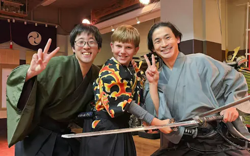Kid posing with a sword at Samurai Experience with two instructors in traditional attire.
