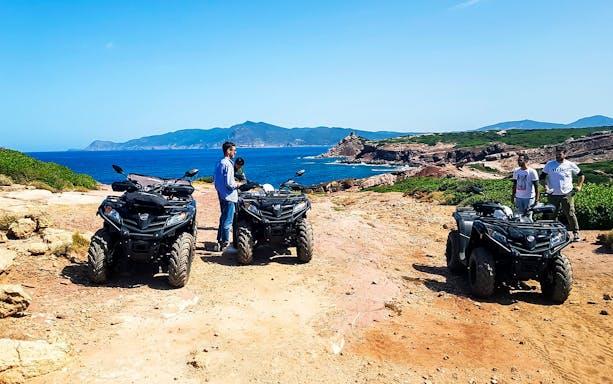 Quad bikers on coastal trail in Alghero, Sardinia with sea and cliffs in background.